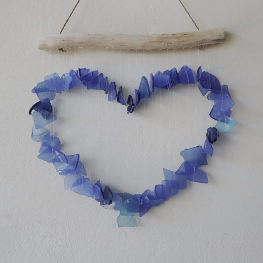 A driftwood-hung mobile, shaped like a heart, is composed of numerous small, translucent blue glass pieces.