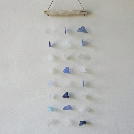 A sea glass mobile hangs from a driftwood bar against a light wall. Various shades of blue and white glass pieces dangle at varying lengths.