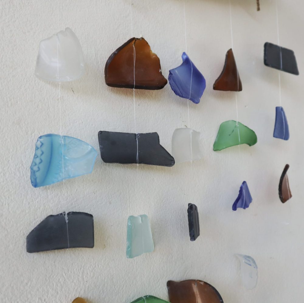 Sea glass pieces hang from thin strings against a white wall, creating a mobile.