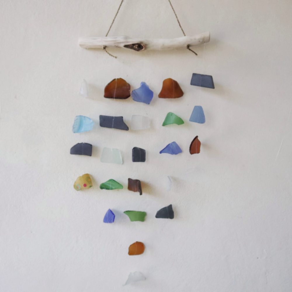 A sea glass mobile hangs against a white wall. Various-colored sea glass pieces are strung and suspended from a driftwood branch.