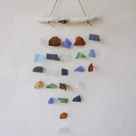 A sea glass mobile hangs against a white wall. Various-colored sea glass pieces are strung and suspended from a driftwood branch.