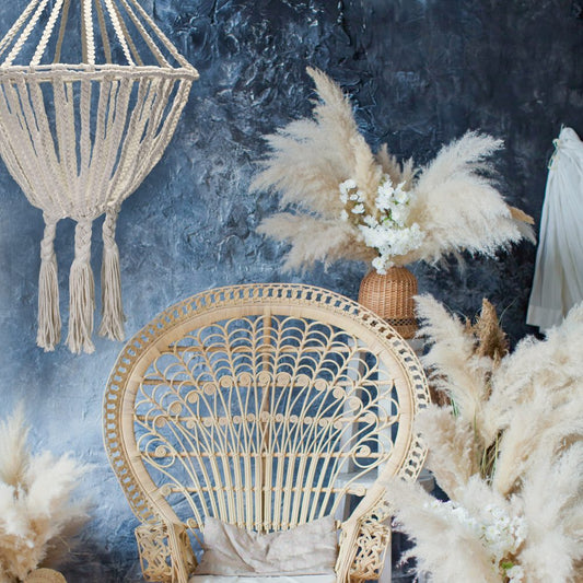 A woven macrame lamp hangs above a wicker peacock chair, which sits amongst pampas grass arrangements against a textured blue wall.