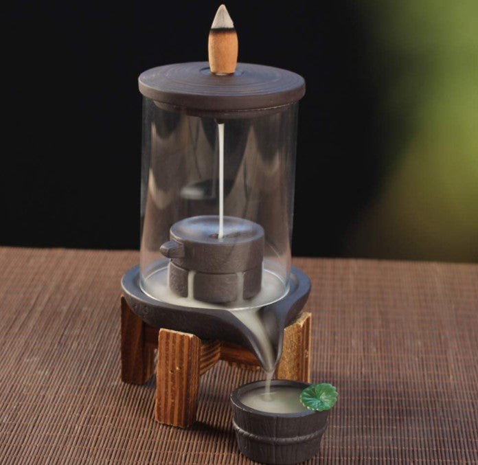 A tabletop water fountain with a cylindrical glass cover and a cone-shaped top discharges water into a small bowl, set upon a wooden stand, against a dark backdrop.