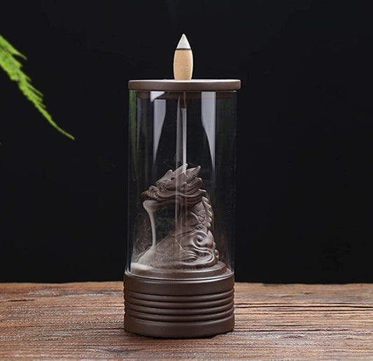 A dragon-shaped incense burner, inside a glass cylinder, emits smoke that cascades downwards. It sits on a wooden surface with a dark, blurred background and a green leaf partially visible.