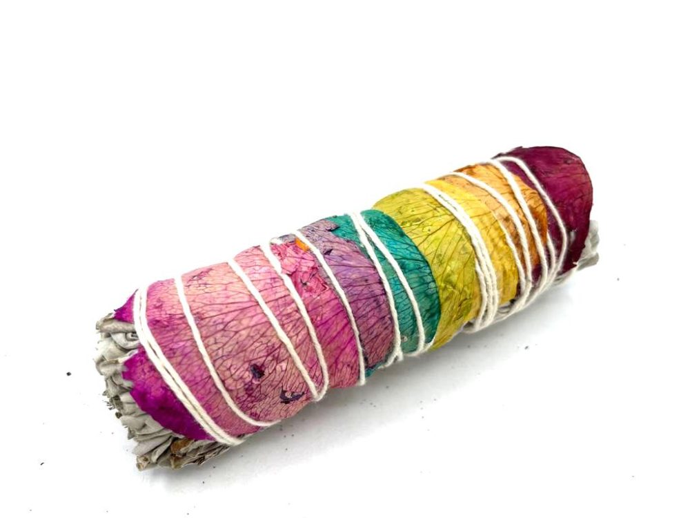 A sage bundle, wrapped in colorful flowers and tied with white string, sits diagonally on a solid white surface. The flowers create a rainbow effect.