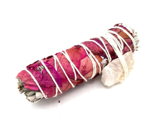 A sage smudge stick, bound with twine and adorned with dried pink petals and a rose quartz crystal, rests on a white surface.