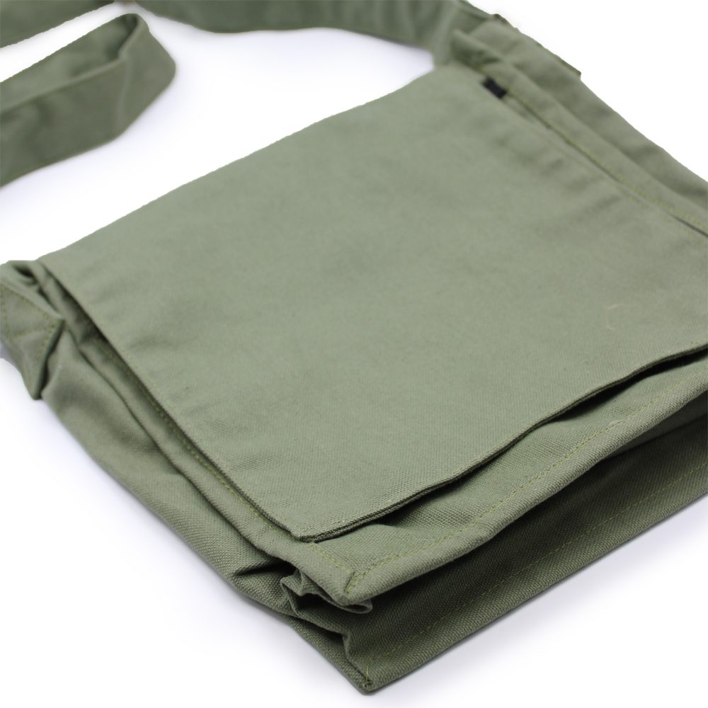 A square, light-green canvas bag with a shoulder strap sits on a white surface. The bag is folded over itself.