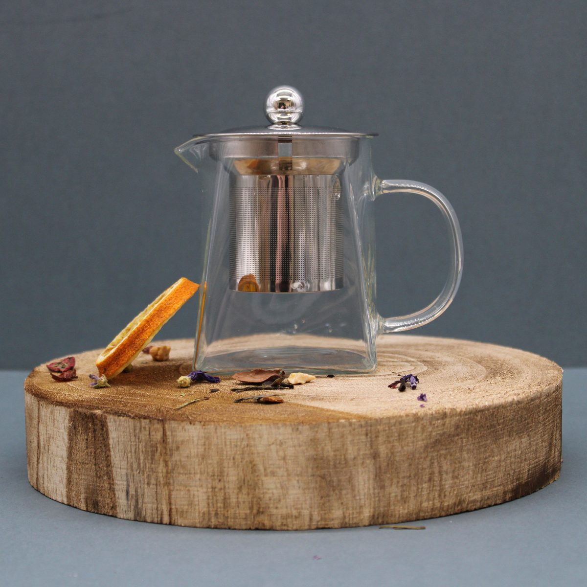 A glass teapot with a metal infuser and lid sits centered on a round wooden platform. Surrounding it are scattered dried herbs and a slice of orange, against a plain backdrop.