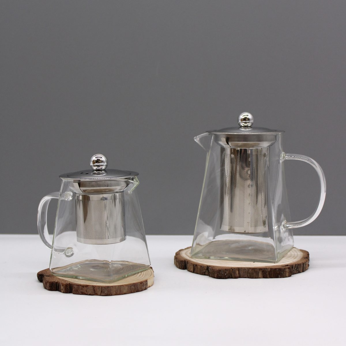 Two glass teapots with metal lids sit on wooden coasters; one is larger than the other, both containing metal infusers, against a gray background on a white surface.