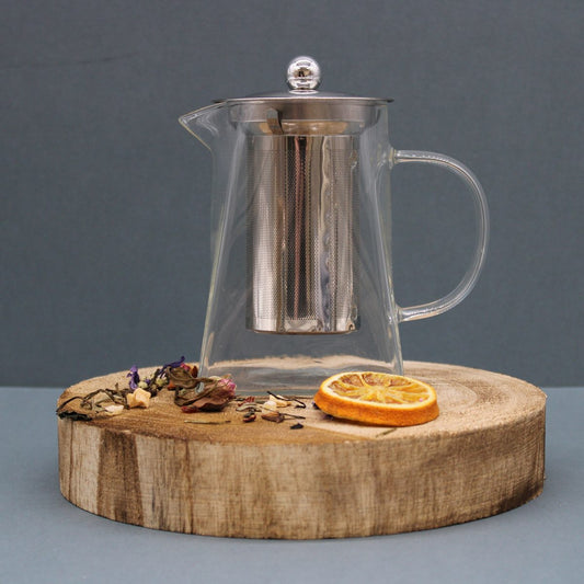 A clear glass teapot with a metal infuser sits on a round wooden base, surrounded by dried herbs and a slice of dried orange, against a neutral background.