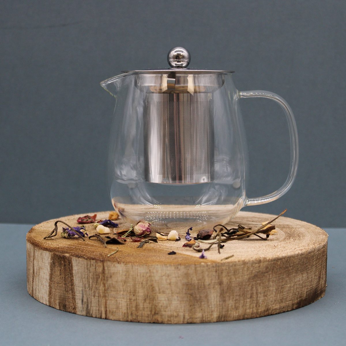 A transparent glass teapot with a metal infuser and lid sits on a wooden slab surrounded by dried herbs and flowers against a plain gray background.