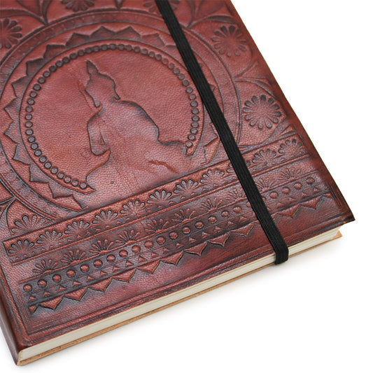 An embossed, leather-bound journal with a black elastic closure rests diagonally, revealing its textured cover featuring a central Buddha figure surrounded by concentric patterns, against a plain white backdrop.