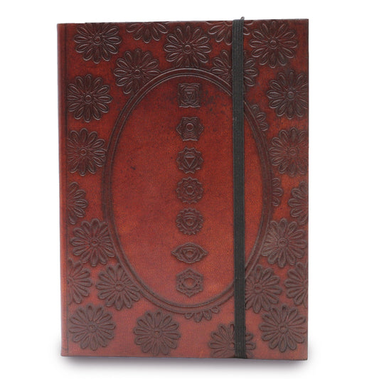 A leather journal, bound by a black elastic strap, displays embossed flowers around an oval. The oval contains embossed spiritual symbols stacked vertically on the cover.