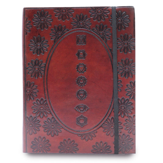 The closed journal, featuring daisy patterns and an oval displaying seven chakra symbols, lays on a white surface. A black elastic band keeps the journal closed.