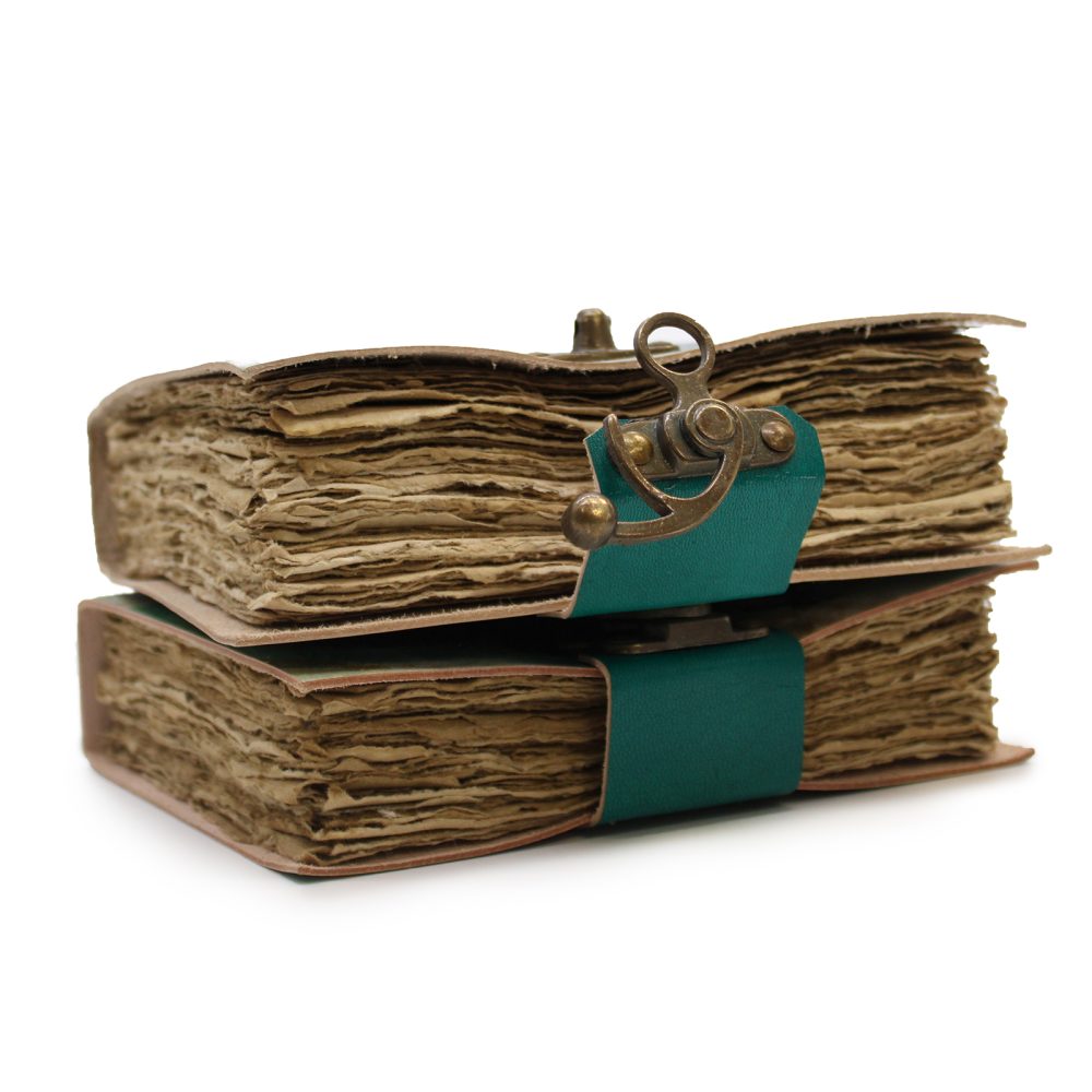 Two aged books sit stacked, closed with a green leather strap and brass clasp. The books have weathered pages and covers, set against a plain white background.