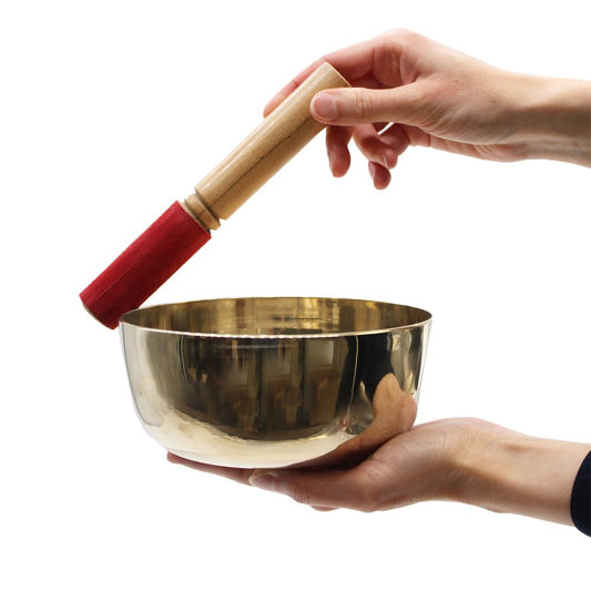 A person holds a Tibetan singing bowl in one hand and a mallet in the other, poised to strike. The background is plain white, focusing on the objects.