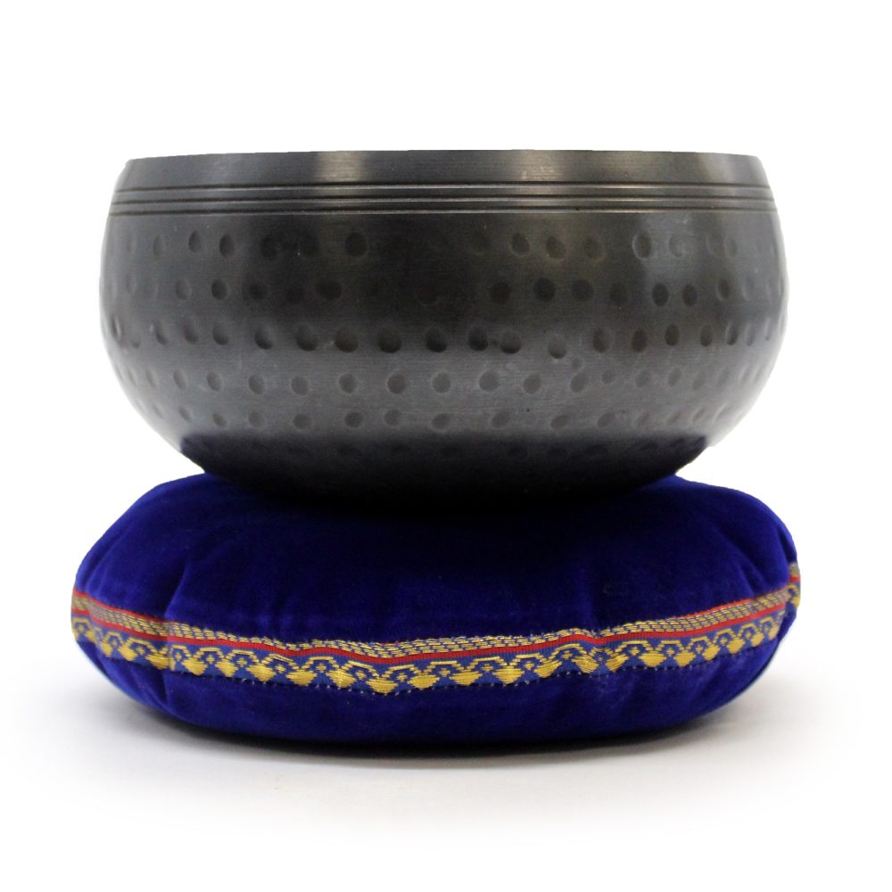 A metallic singing bowl, featuring a dotted pattern, rests on a deep blue velvet cushion with an ornate gold and red trim, set against a plain white background.