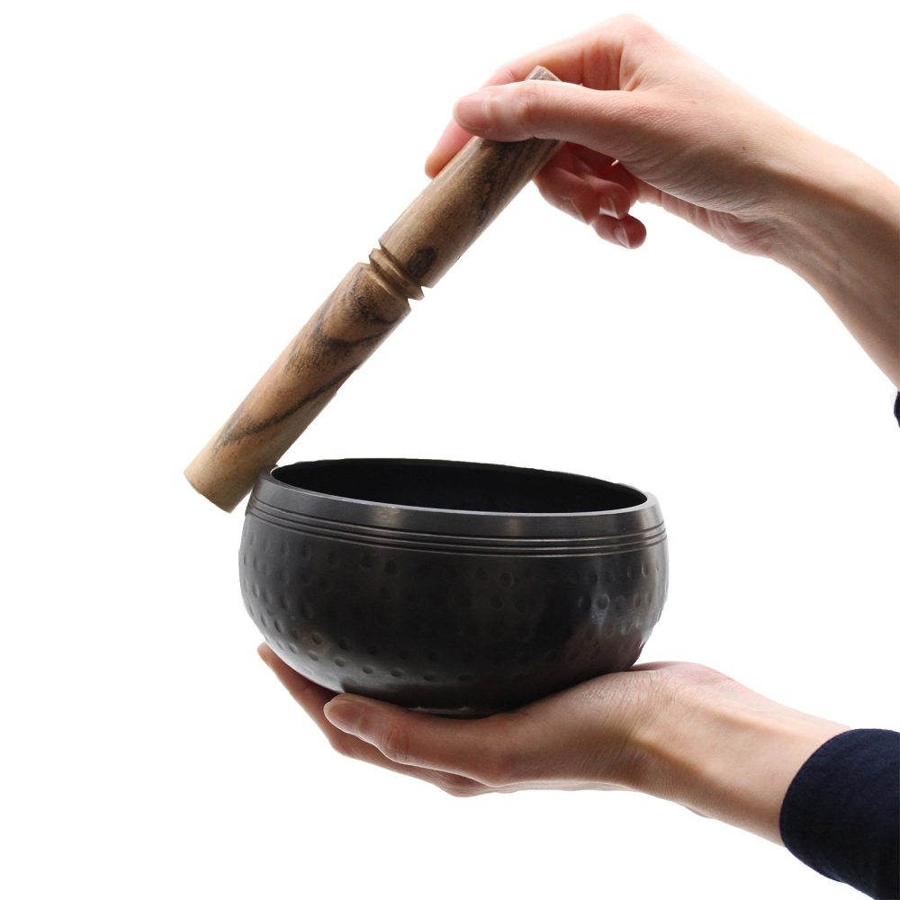 A dark, textured singing bowl is held in one hand, while the other hand uses a wooden mallet to play it, isolated against a plain white background.