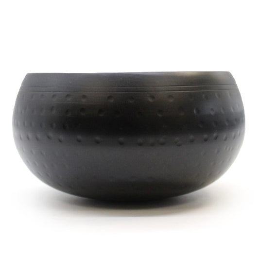 A black, round metal bowl sits on a plain white background. The bowl features a textured surface with small indentations evenly distributed across its exterior.