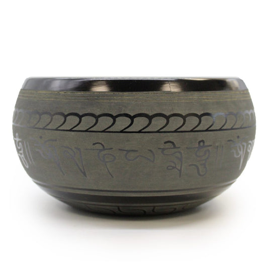 A metallic singing bowl with intricate carvings and Sanskrit script encircling its exterior; it rests against a plain white background.