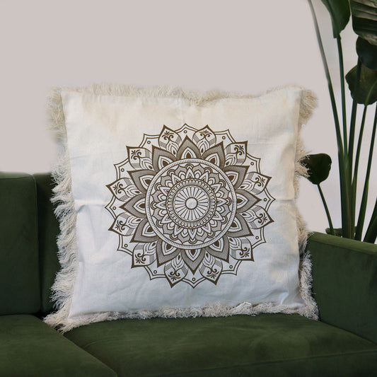 A beige square cushion, featuring a dark-brown mandala print and frayed edges, rests on a dark-green sofa.