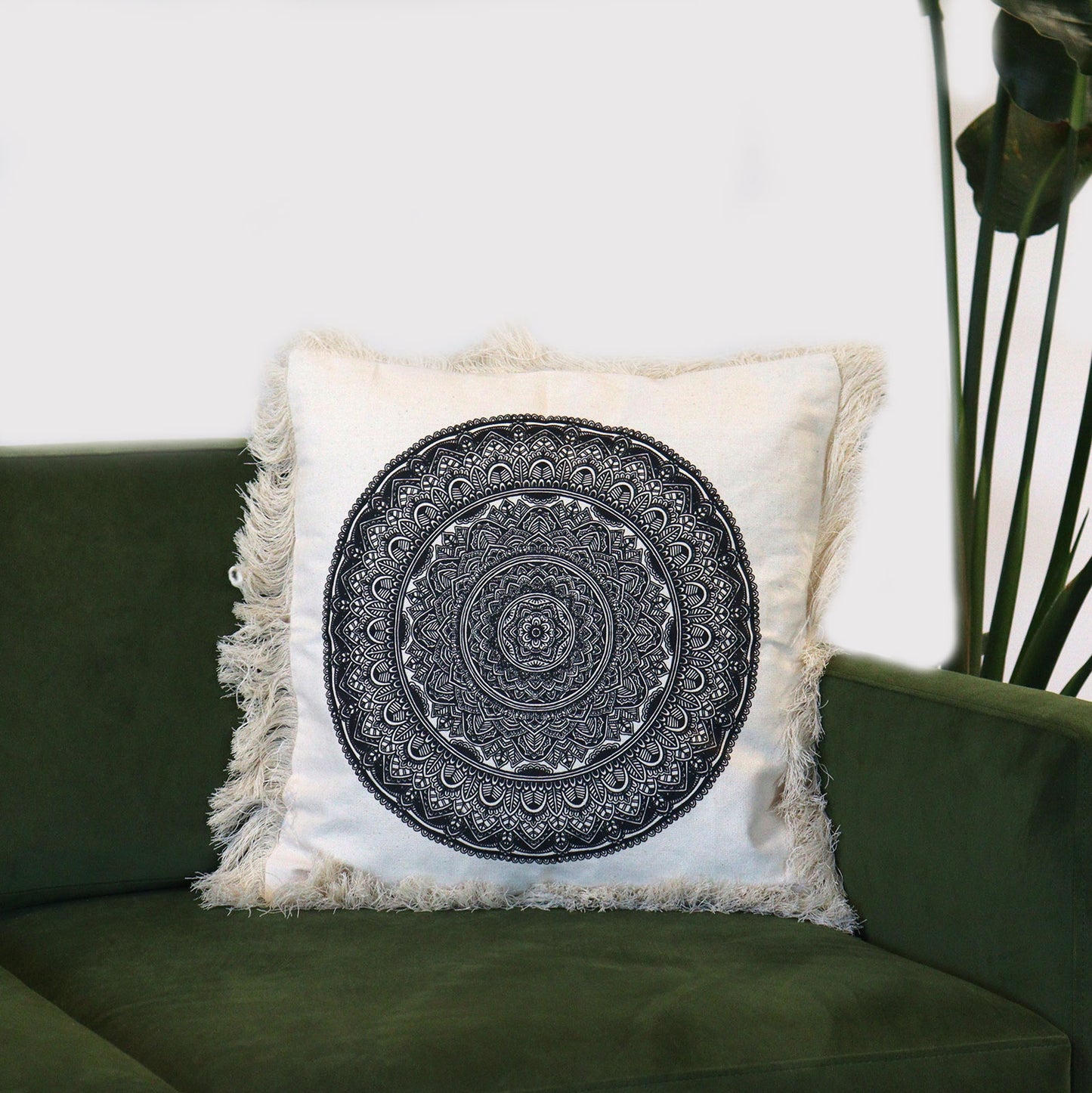 A fringed cushion, featuring a central black mandala design, rests on a dark-green sofa.