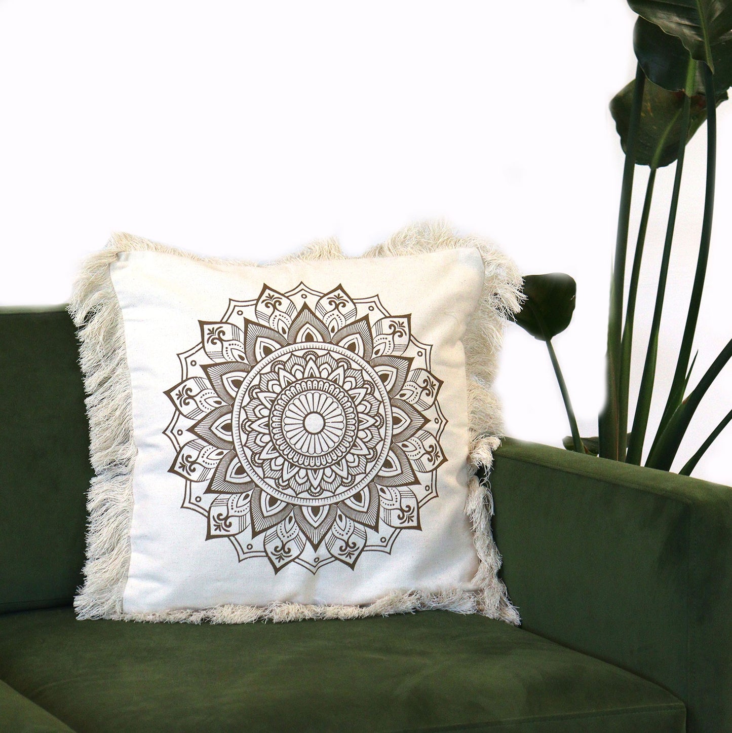 A fringed off-white cushion, featuring a brown mandala print, rests on a dark-green couch. A plant stands nearby.