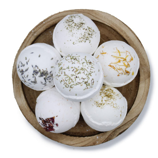 Six white bath bombs, decorated with dried herbs and petals, are arranged in a wooden bowl, creating a natural, aromatic display.
