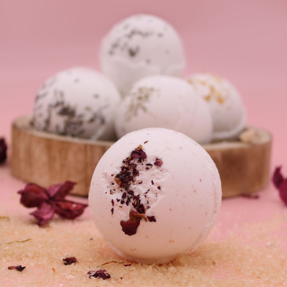 A bath bomb with dried petals sits on pink granules, surrounded by similar bath bombs in a wooden bowl against a pink background.