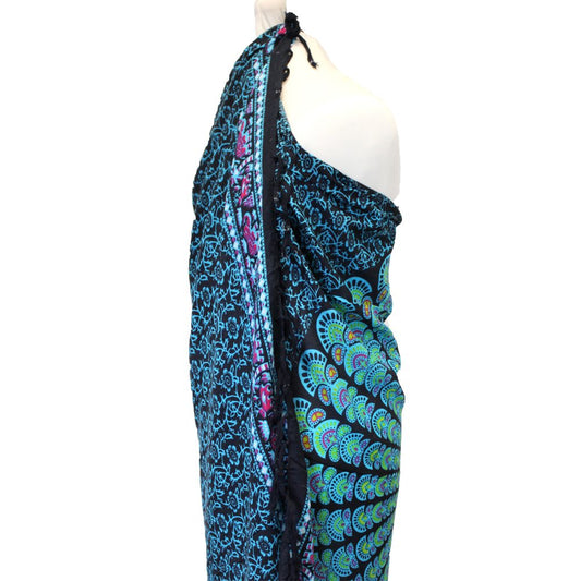 A sarong, draped on a mannequin, displays a teal and black mandala pattern. The sarong features fringed edges. The background is white.