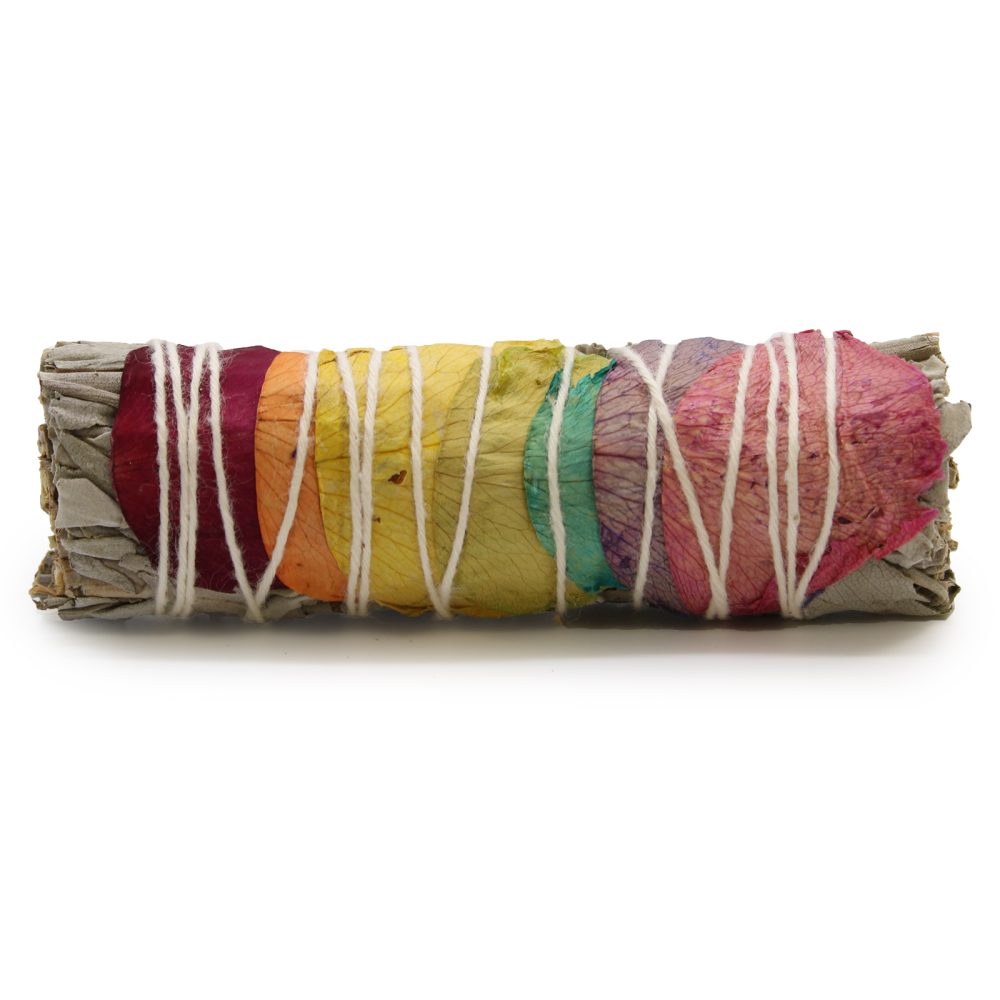 A sage bundle, secured with white string, has colorful flower petals arranged in a rainbow pattern. It sits on a white surface.