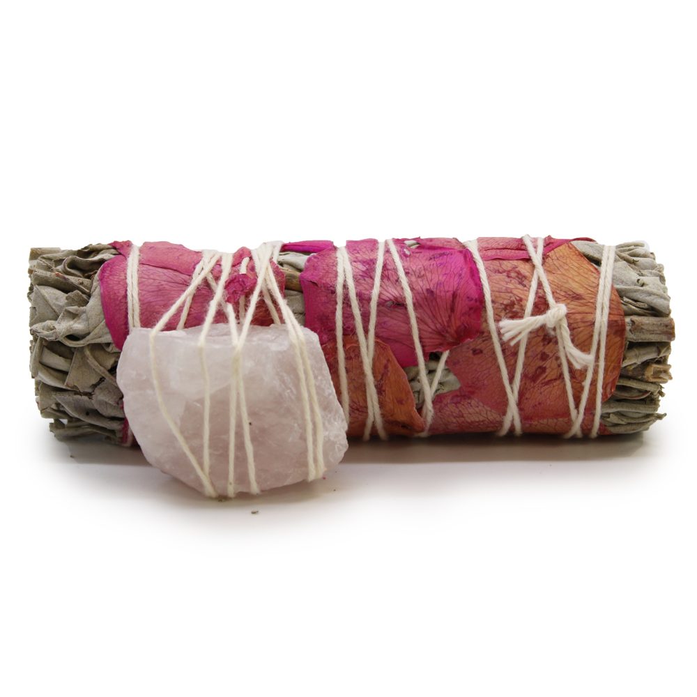 Bundle of sage and rose petals are tied together with white string, with a rose quartz crystal attached. The objects lay on a white surface.