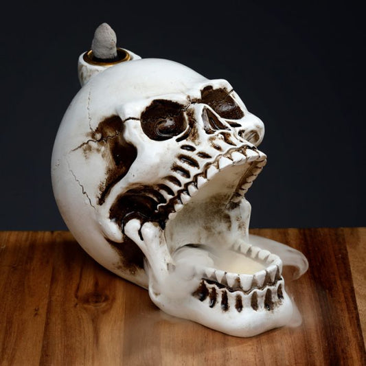 A ceramic skull incense holder emits smoke from its open mouth, sitting on a wooden surface against a dark background.