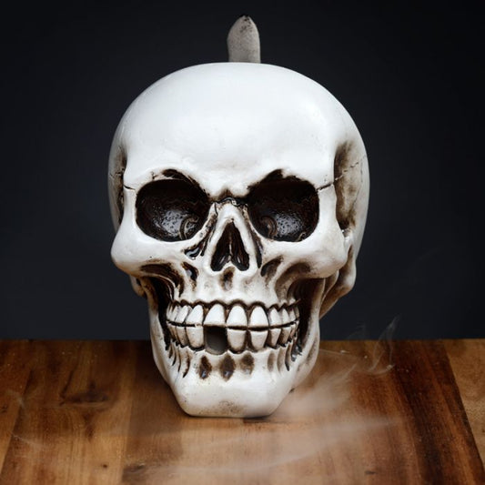 A ceramic skull candle emits smoke on a wooden surface. It has a realistic design with prominent teeth and eye sockets, creating an eerie atmosphere against a dark background.