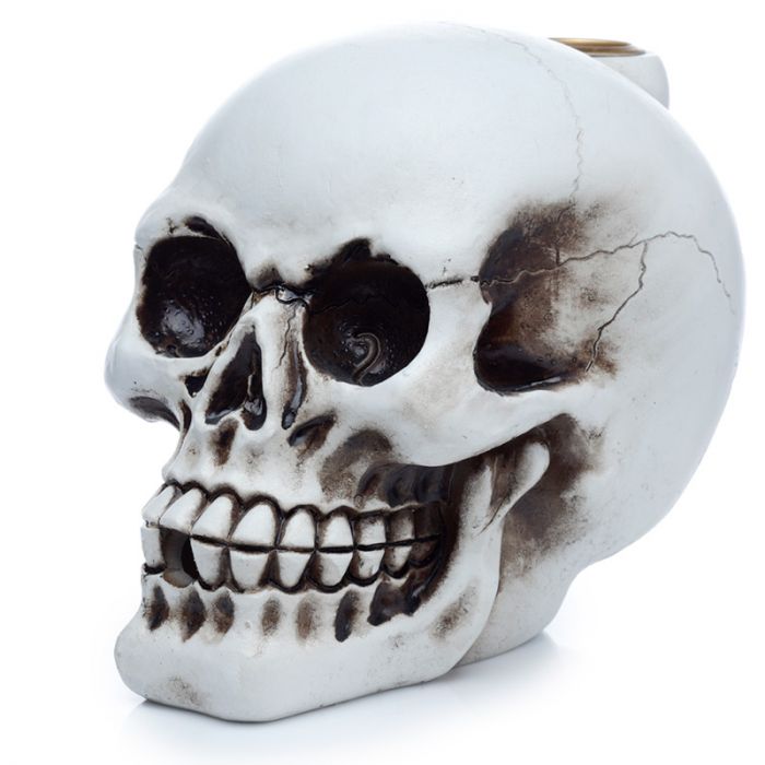 A detailed ceramic skull figurine rests on a white background, showing distinct eye sockets and teeth, with dark shading and a subtle crack for added realism.