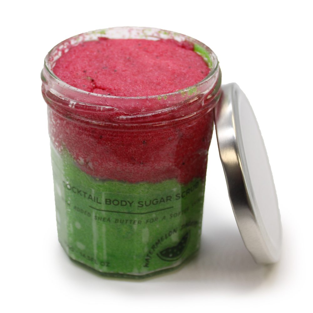 Open glass jar holds pink and green body sugar scrub and its lid is placed next to it. The jar reads "COCKTAIL BODY SUGAR SCRUB ADDED SHEA BUTTER FOR A SOFTER SMOOTHER SKIN WATERMELON DREAM".