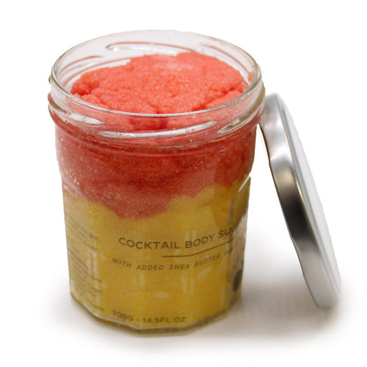 Glass jar containing pink and yellow layered body scrub with its metal lid leaning beside it on a white surface; label text reads "COCKTAIL BODY SUGAR", "WITH ADDED SHEA BUTTER", "300G - 14.5FL OZ".