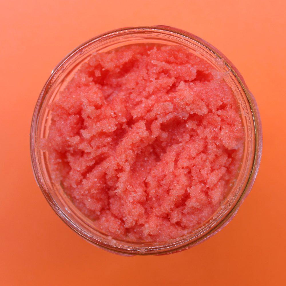 A glass jar is filled with pink scrub. The jar is set on an orange background.