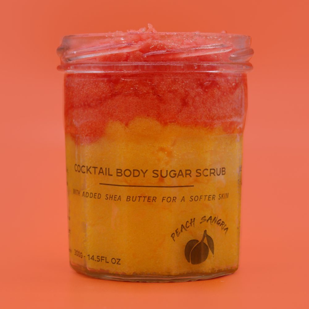 A jar filled with a layered, gradient body sugar scrub sits against a peach-colored background. Text on the jar reads, "COCKTAIL BODY SUGAR SCRUB WITH ADDED SHEA BUTTER FOR A SOFTER SKIN PEACH SANGRIA 300G-14.5FL OZ."