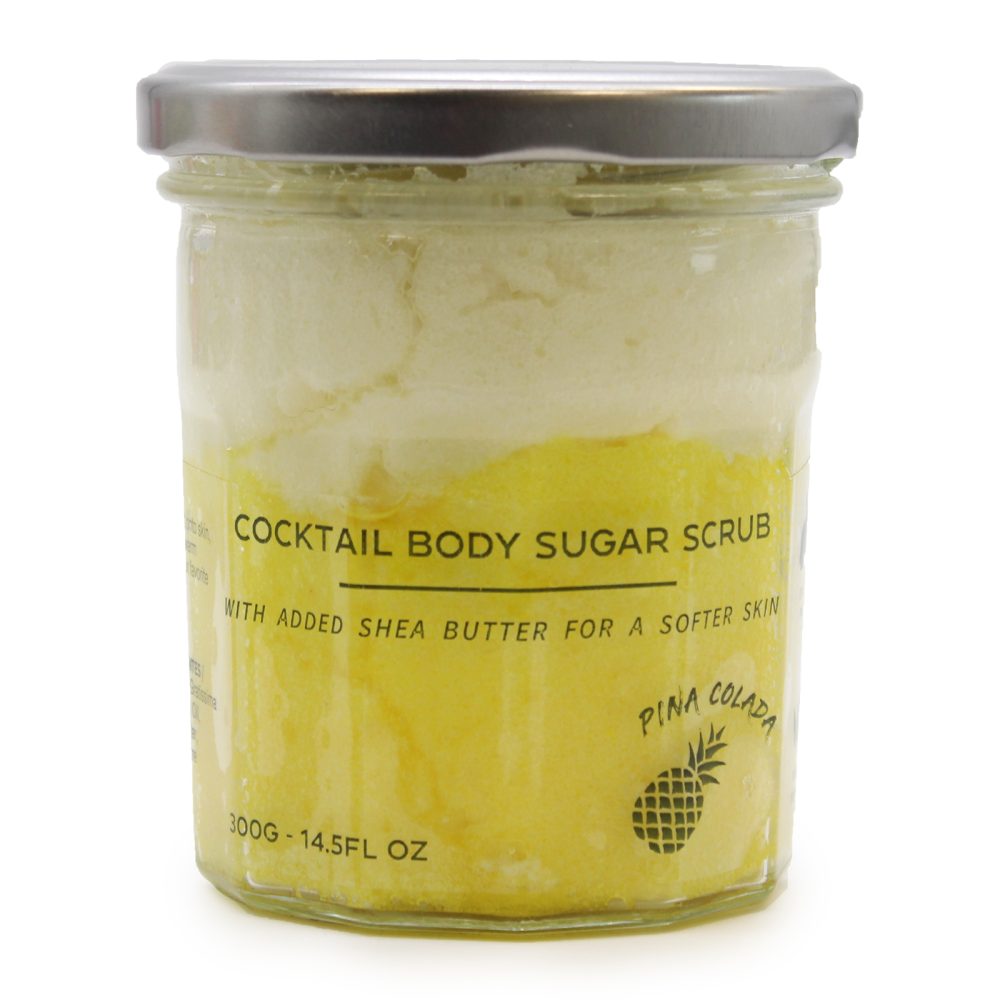 A sealed glass jar displays "COCKTAIL BODY SUGAR SCRUB/WITH ADDED SHEA BUTTER FOR A SOFTER SKIN/PINA COLADA" labels over a layered white-to-yellow scrub, on a white surface. The jar is capped with a metal lid and weighs "300G-14.5FL OZ".