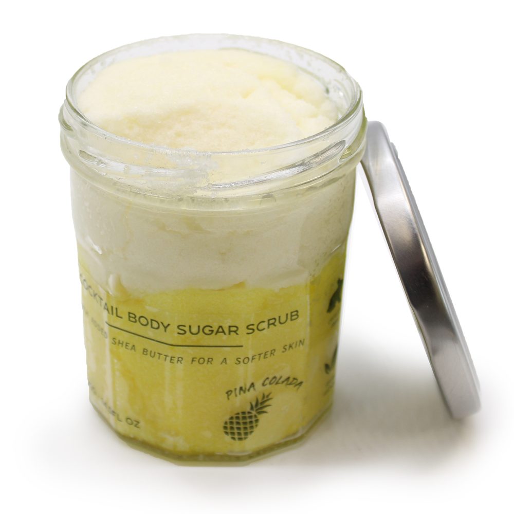 Open glass jar displays layered "Cocktail Body Sugar Scrub" with text stating "... shea butter for a softer skin, Pina Colada" and a pineapple design on a white surface.