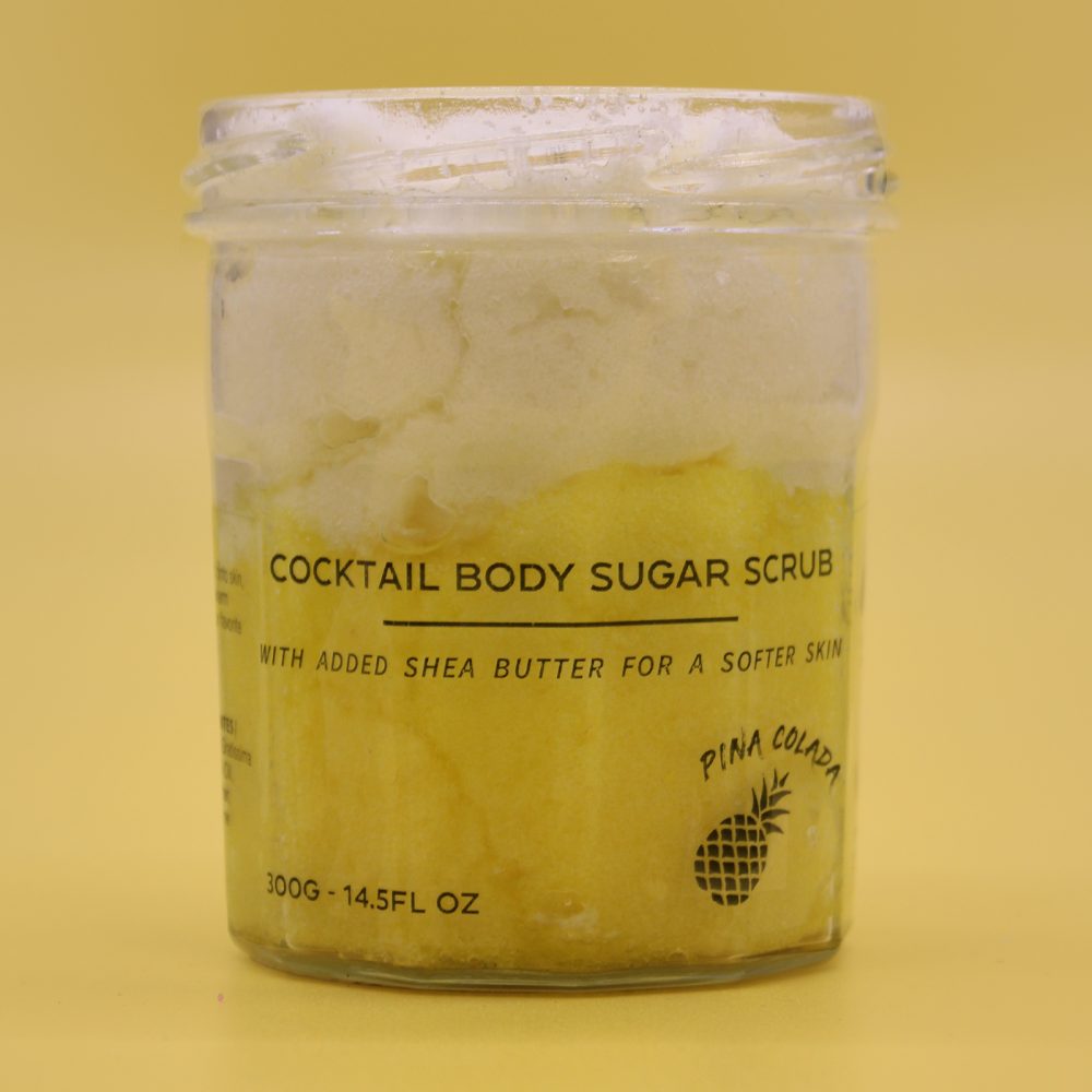 Body scrub sits inside a clear jar. It is layered yellow and white, and it sits on a pale yellow background. Text on the front reads "COCKTAIL BODY SUGAR SCRUB, WITH ADDED SHEA BUTTER FOR A SOFTER SKIN, PINA COLADA, 300G - 14.5FL OZ."