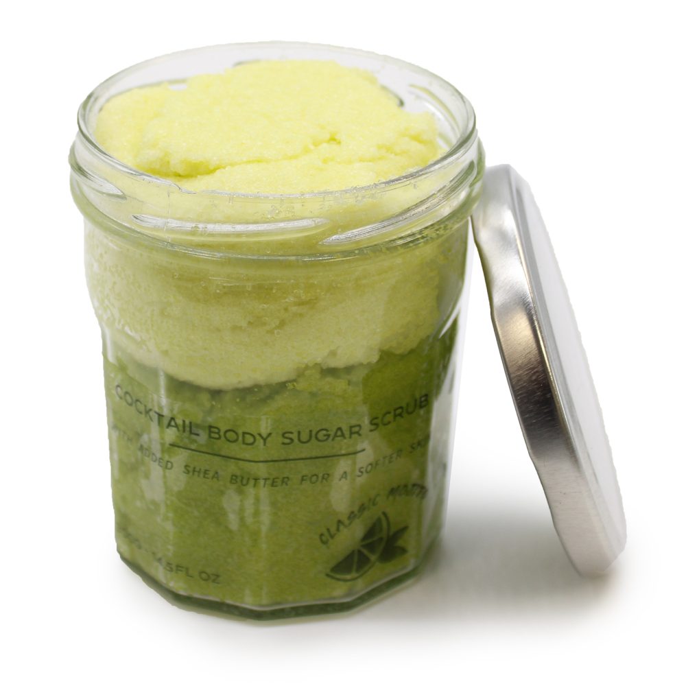 A jar of green-and-yellow body scrub with the words "COCKTAIL BODY SUGAR SCRUB, WITH ADDED SHEA BUTTER FOR A SOFTER SKU, CLASSIC MAD 700-145FL OZ" on its label sits against a white background. The lid is leaning against the side of the jar.