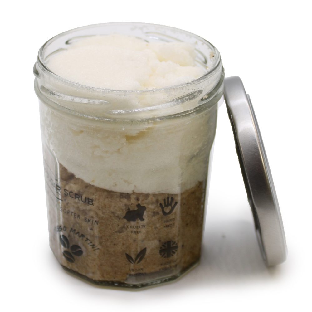A layered scrub, with text reading “SUGAR SCRUB, SOFTER SKIN, XO MARTINI, CRUELTY FREE, HAND MADE, VEGAN, MADE IN ENGLAND” sits in an open glass jar next to its unattached silver lid on a white surface.