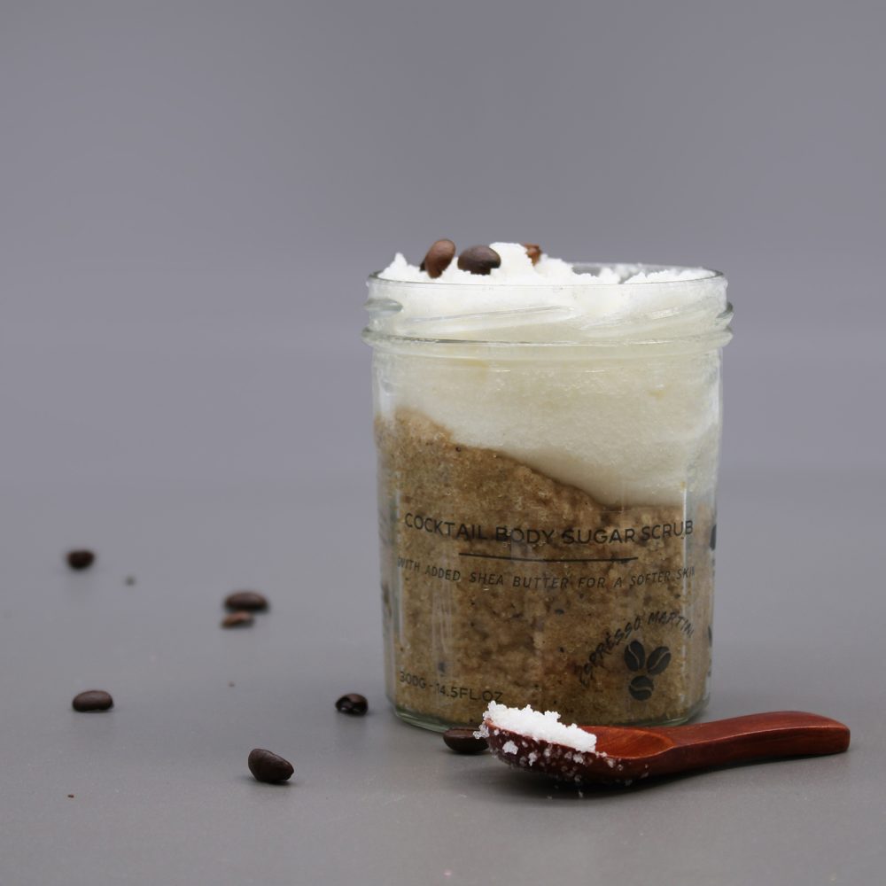 The jar of body scrub sits on a gray surface. The jar has a brown sugar-like substance topped with white cream and scattered coffee beans, with a small wooden spoon filled with the body scrub next to it.
It contains the text "COCKTAIL BODY SUGAR SCRUB WITH ADDED SHEA BUTTER FOR A SOFTER SKI 300G-14.5FL.OZ".