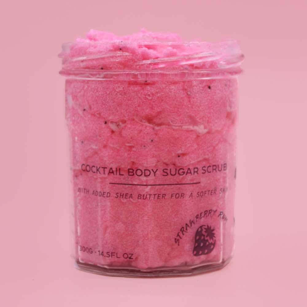 A jar holds pink body scrub. The label reads: "COCKTAIL BODY SUGAR SCRUB WITH ADDED SHEA BUTTER FOR A SOFTER SK," "STRAWBERRY RUM," and "300G-14.5FL OZ". The jar sits on a light pink background.