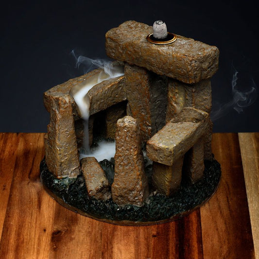 Stone-like incense burner with smoke flowing down creates a waterfall effect on a wooden surface. The structure mimics ancient stone formations, surrounded by a dark backdrop.