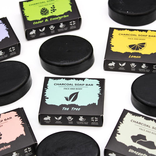 Charcoal soap bars and boxed packages arranged on a white surface, labeled CHARCOAL SOAP BAR FACE AND BODY Tea Tree, CHARCOAL SOAP BAR FACE AND BODY Lemon, CHARCOAL SOAP BAR FACE AND BODY Cedar & Eucalyptus, CHARCOAL SOAP BAR FACE AND BODY Unscented, icons reading CRUELTY FREE VEGAN FRIENDLY HAND MADE PARABEN, SLS & PALM OIL FREE MADE IN ENGLAND