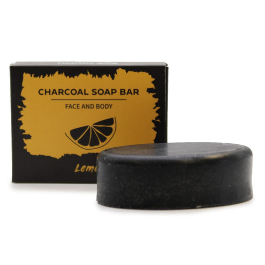 A round black charcoal soap bar rests before its yellow-and-black box on a white surface, labeled CHARCOAL SOAP BAR, FACE AND BODY, Lemon.