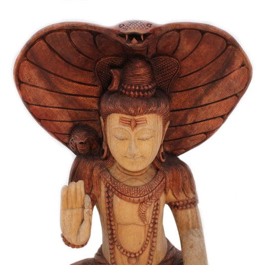 A wooden sculpture depicts a deity sitting with eyes closed, right hand raised in blessing. A hooded cobra shelters the figure, enhancing the serene and spiritual atmosphere.